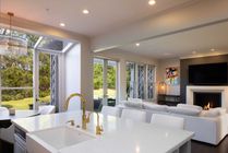 A bright, modern open-plan kitchen and living room featuring a white island, gold faucet, and a fireplace with a TV.