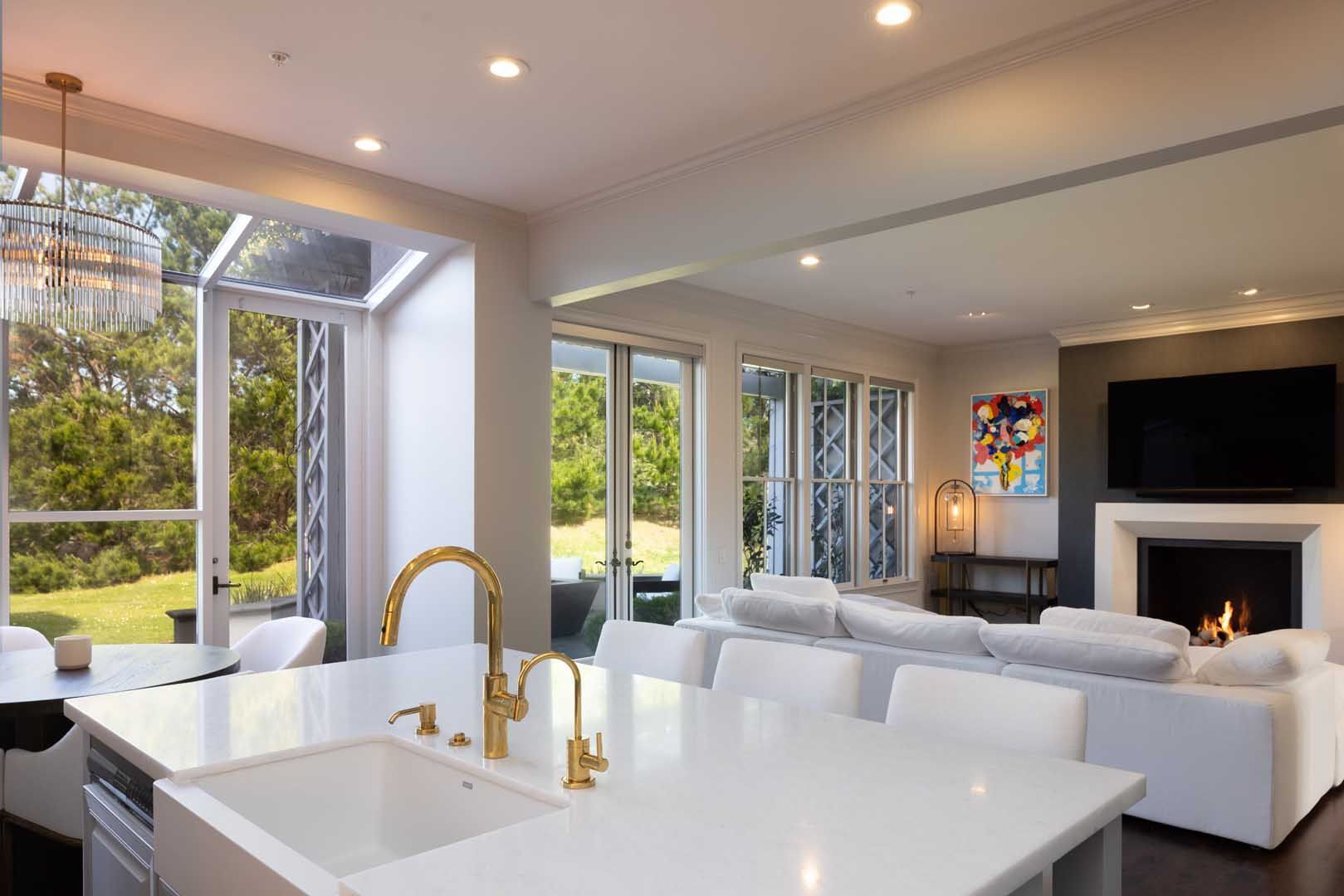 A bright, modern open-plan kitchen and living room featuring a white island, gold faucet, and a fireplace with a TV.