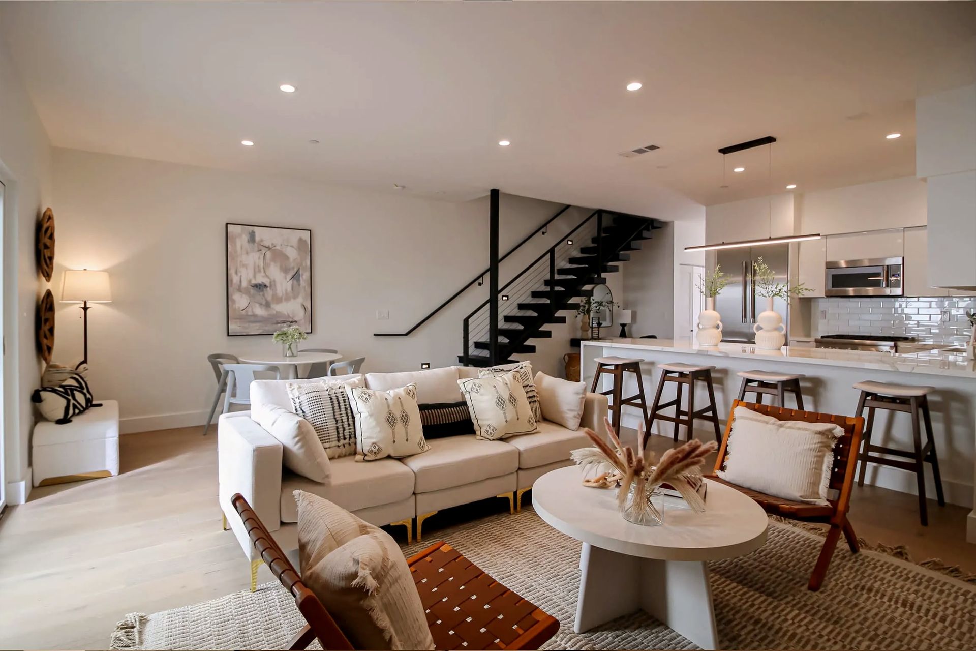 A modern open-concept home interior featuring a neutral-toned living room, dining area, and kitchen with a dark staircase.
