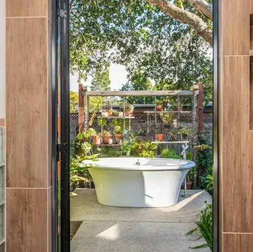 A freestanding white bathtub sits on a patio surrounded by plants, viewed through an open doorway framed in wood tile.