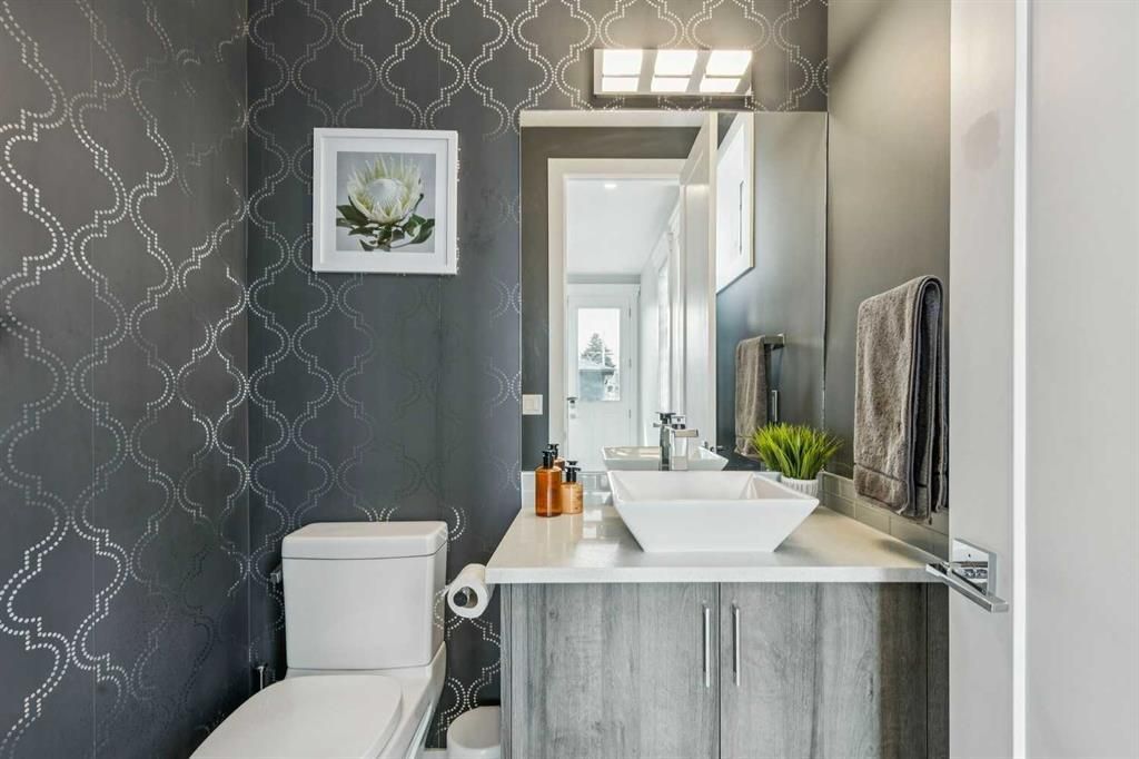 Narrow gray bathroom with patterned wallpaper, vanity sink, toilet, and mirror lights