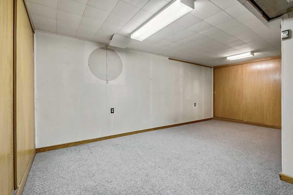 Empty room with white walls, wood-paneled corner, and gray carpeted floor under fluorescent ceiling lights