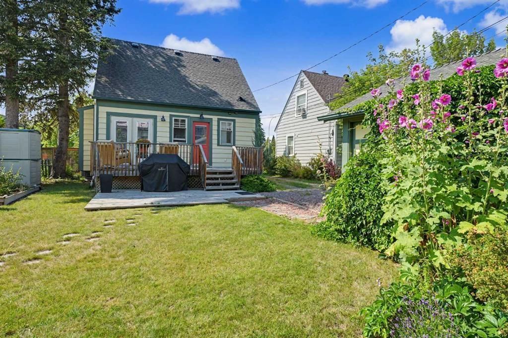 Small cottage with porch in a sunny yard, surrounded by grass, trees, and pink flowers