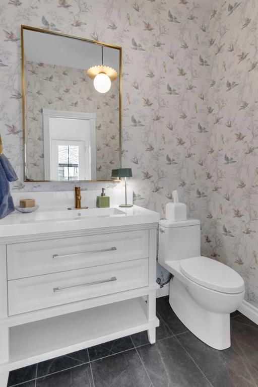 Bright bathroom with white vanity, gold-framed mirror, lit wall sconce, and toilet beside floral wallpaper