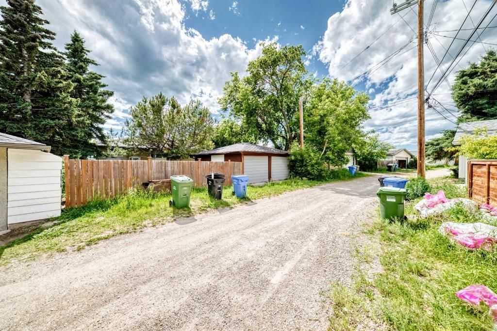 Gravel alley beside wooden fences and sheds, with trash bins, trees, and pink flowers under a cloudy sky