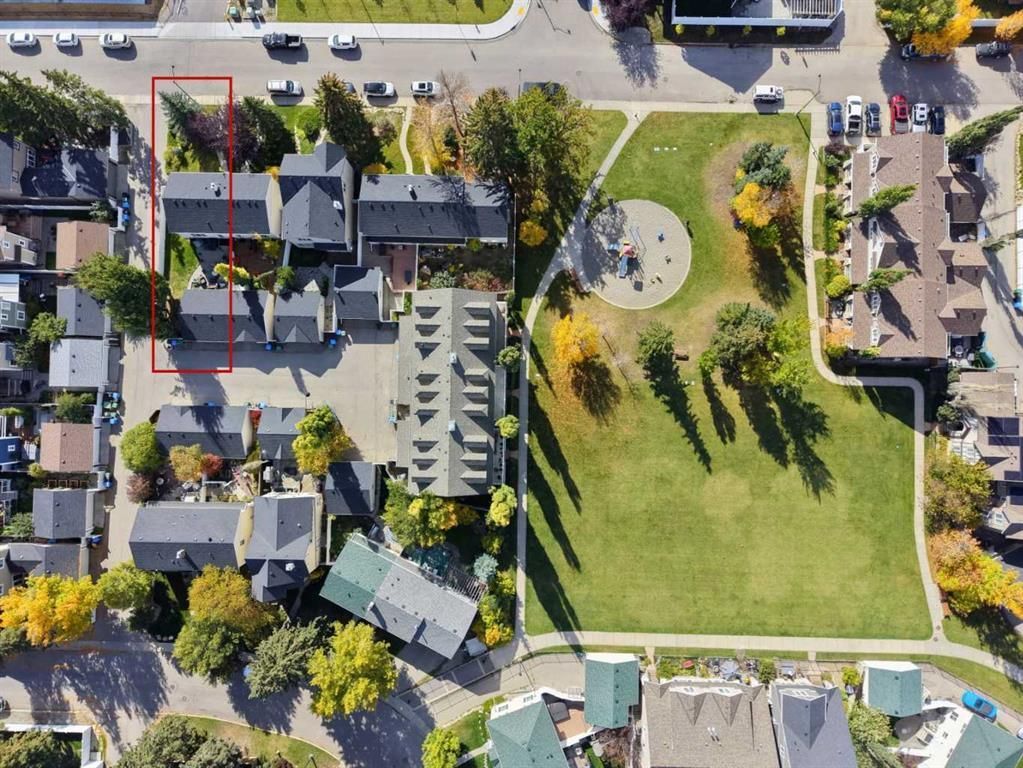 Aerial view of a park with a large lawn, trees, paths, and nearby buildings and parked cars.