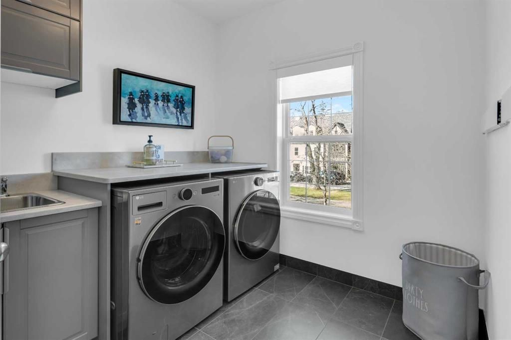 Modern laundry room with front-loading washer and dryer, gray countertops, sink, window, and trash can.