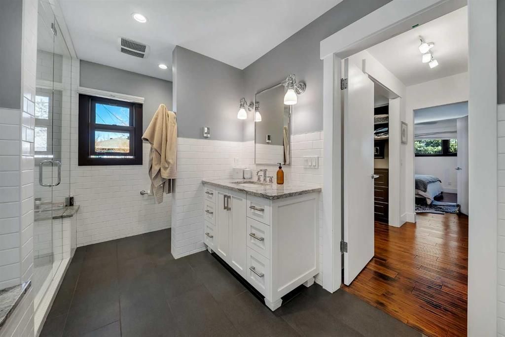 Bright bathroom with white vanity, tiled shower, and dark floor; open doorway shows hardwood hallway.