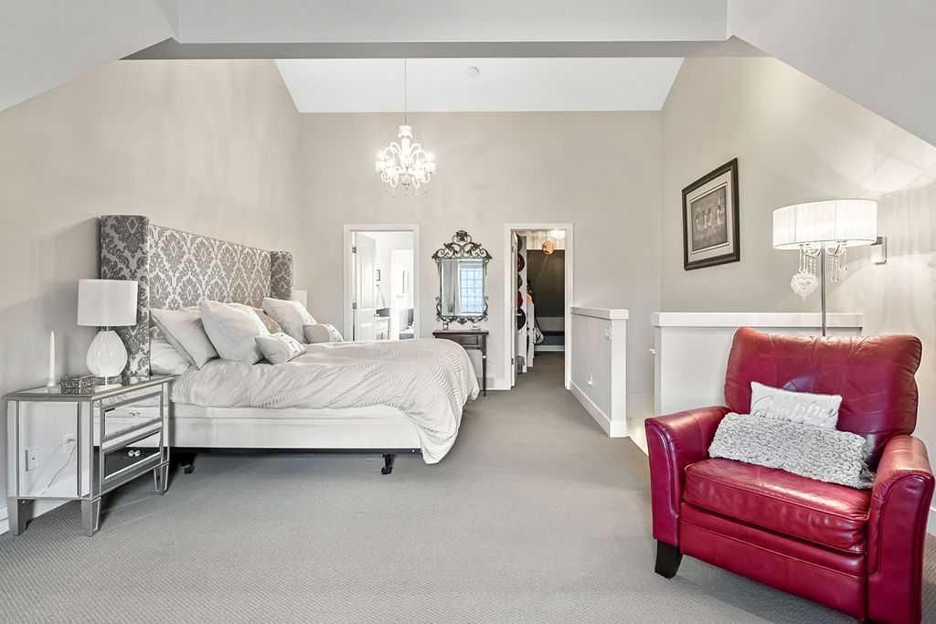 Elegant white bedroom with chandelier, mirrored vanity, and a bright red armchair on gray carpet