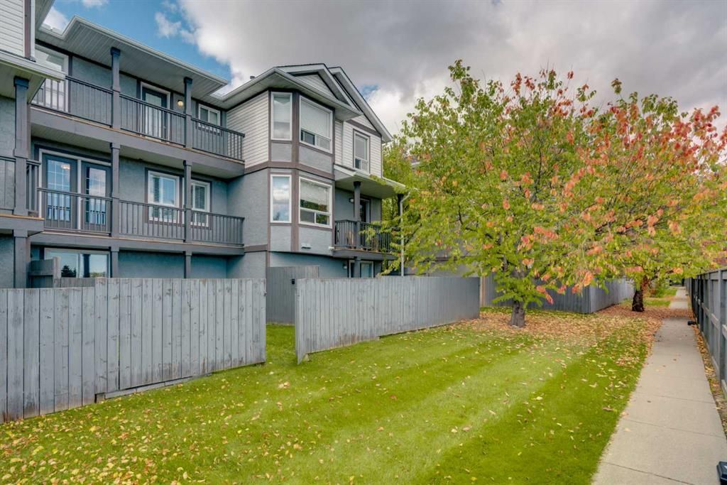 Apartment building behind a wooden fence, with green lawn, sidewalk, and a tree with autumn leaves.