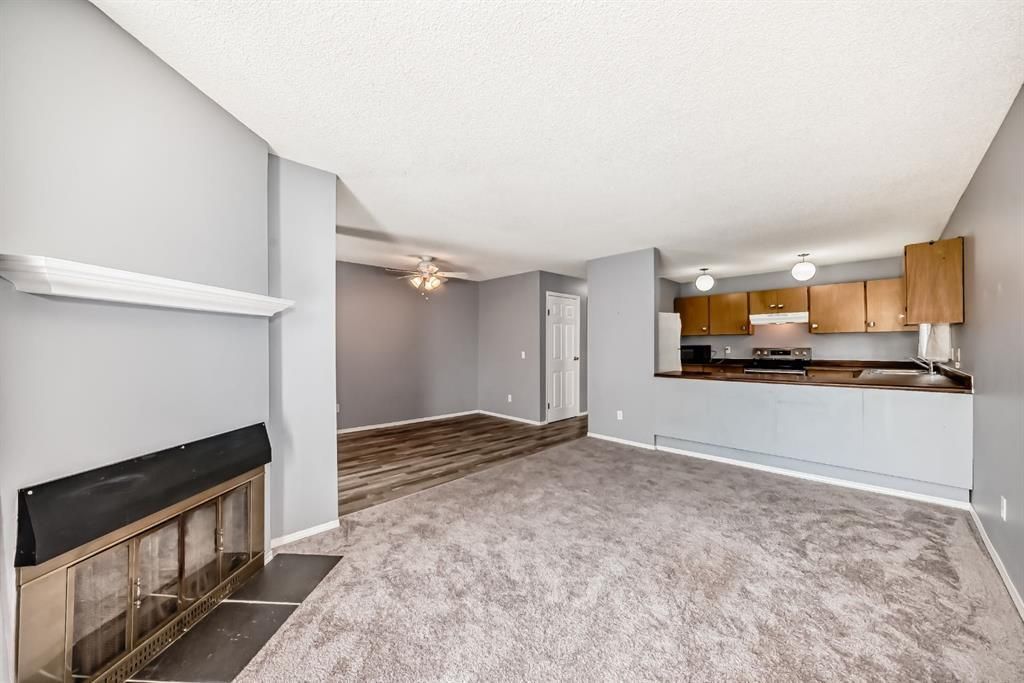 Empty living room with fireplace, gray carpet, and open view to a kitchen with wooden cabinets.