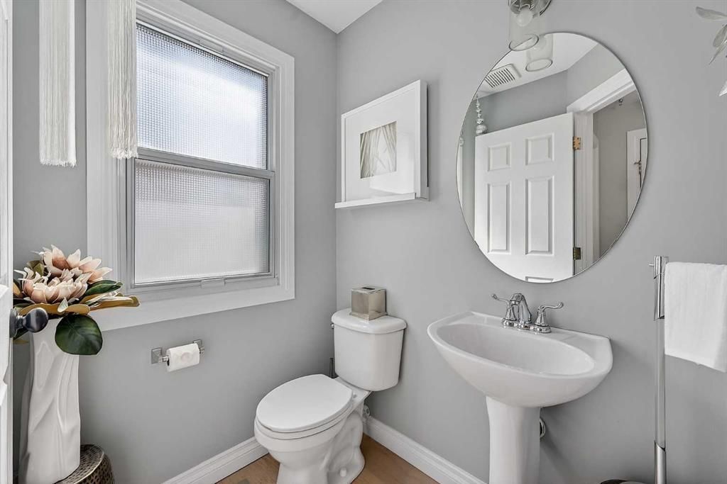 Bright gray bathroom with toilet, sink, round mirror, window, and floral vase on a small shelf.