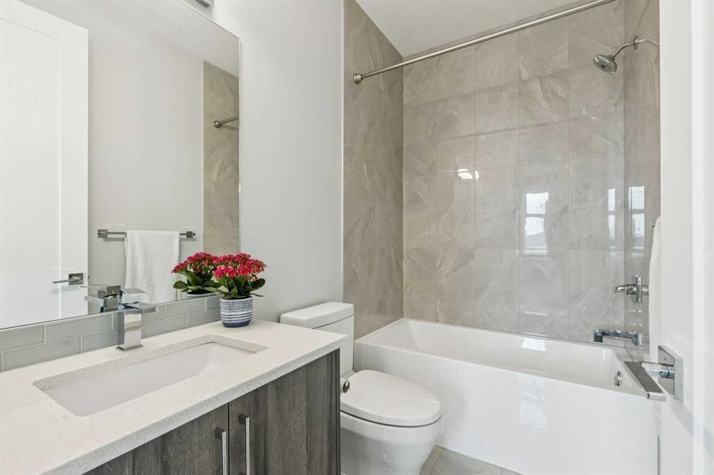 Bright modern bathroom with white vanity, flowers, toilet, and tiled bathtub shower area.