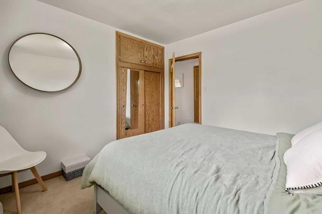 Minimal bedroom with a bed, round mirror, wooden door, and chair on a neutral wall.
