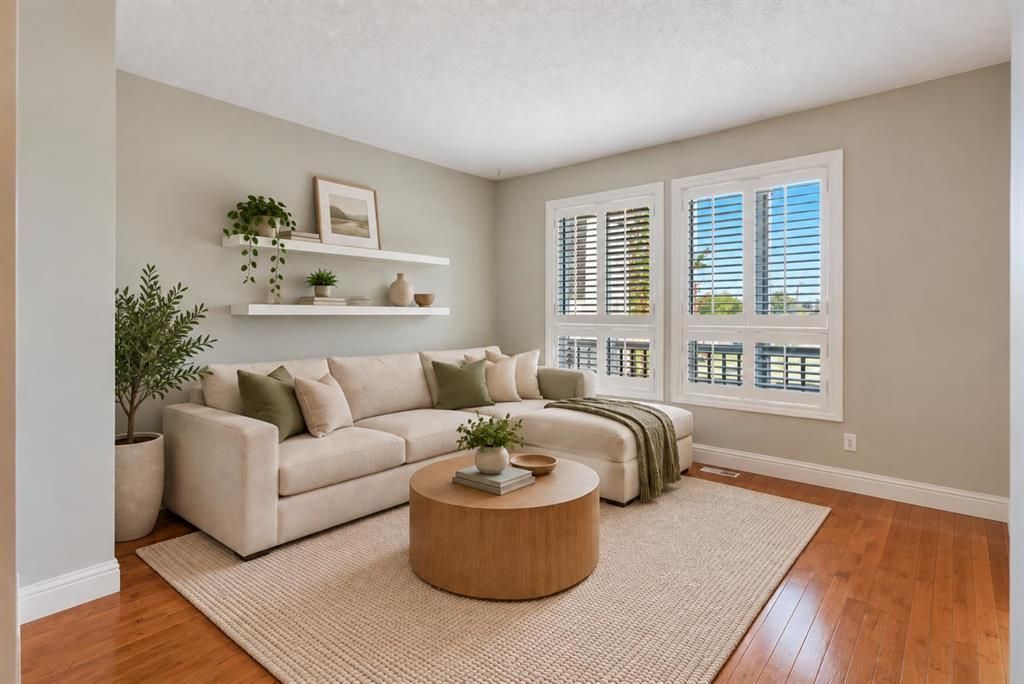 Bright living room with beige sofa, round wood table, wall shelves, and large windows with shutters