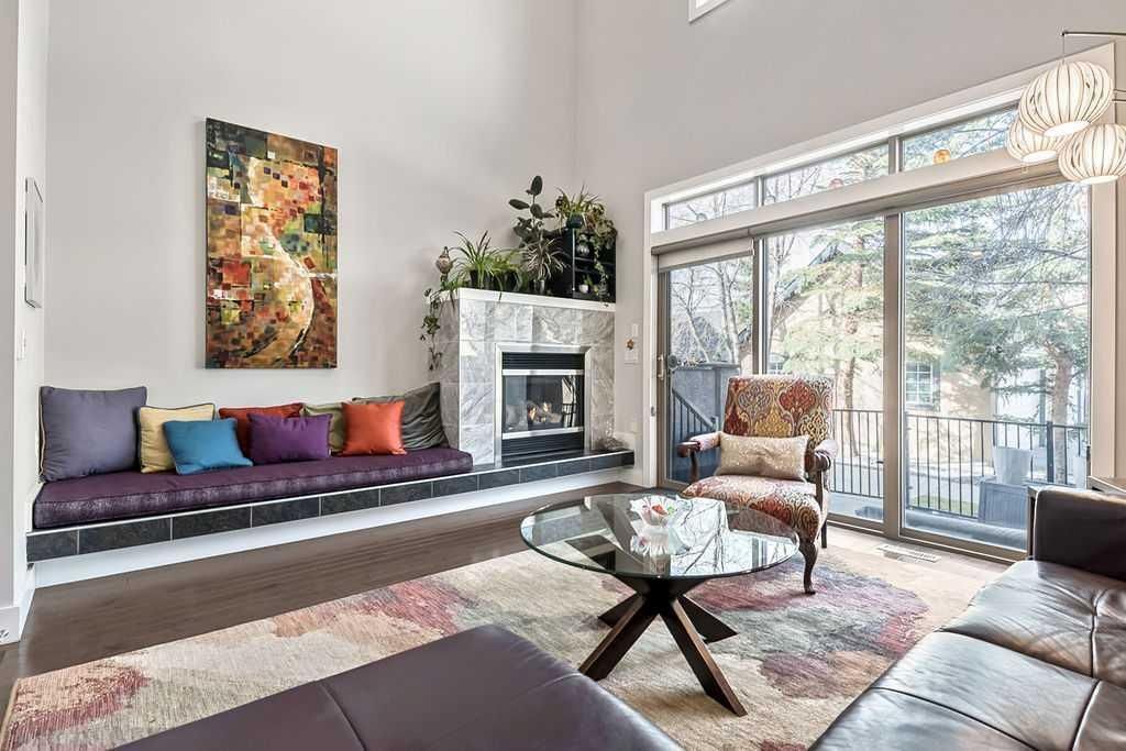 Modern living room with sectional sofa, fireplace, colorful wall art, glass coffee table, and large windows.