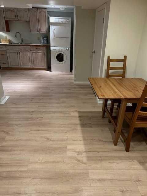 Empty apartment with light wood flooring, small dining table and chairs, and a kitchen in the background