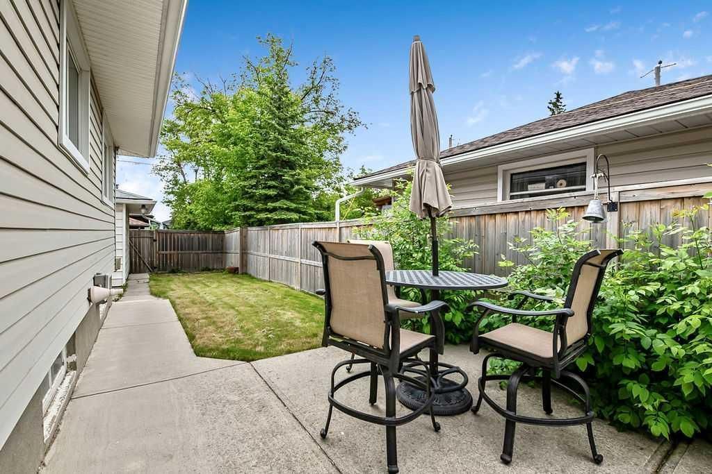 Patio with two chairs and table beside a house, surrounded by a fenced backyard and greenery.