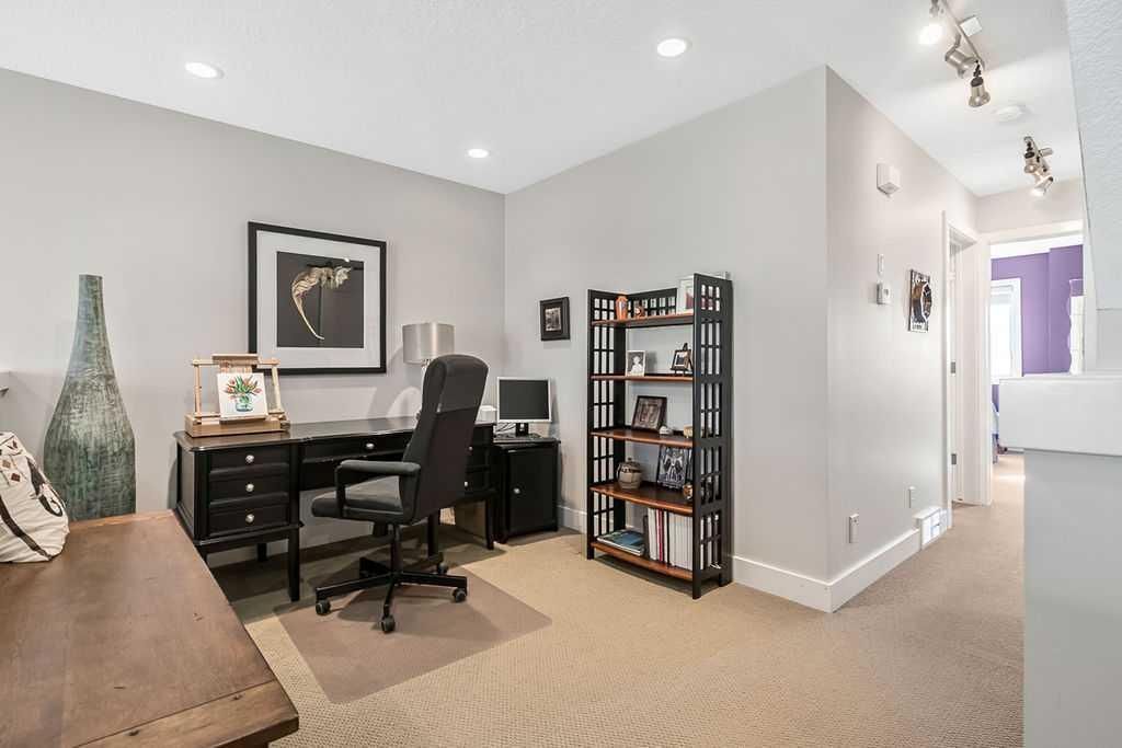 Modern home office with black desk, chair, bookshelf, and framed wall art in a carpeted hallway area