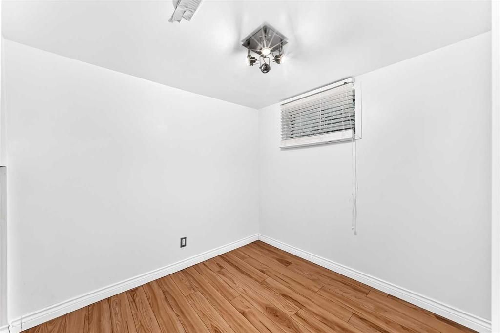 Empty white room with wood floor, ceiling light, and a small window with blinds