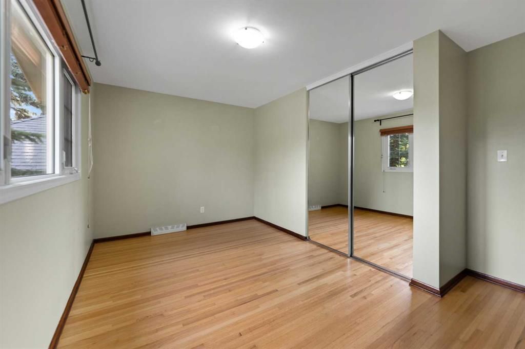 Empty bedroom with wood floors, beige walls, a window, and mirrored closet doors