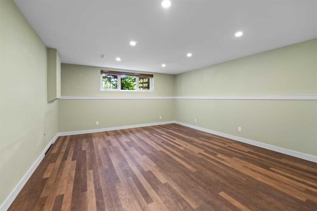 Empty beige room with wood floors, recessed lights, and a small window on the far wall