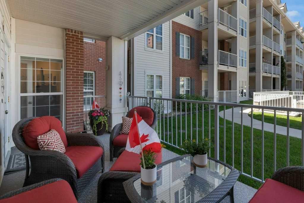 Covered apartment balcony with red-cushioned chairs, glass table, and view of nearby buildings and lawn
