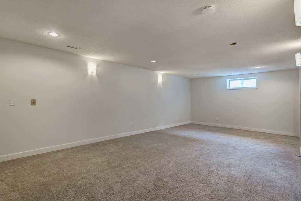 Empty finished basement with beige carpet, white walls, recessed lights, and a small window.