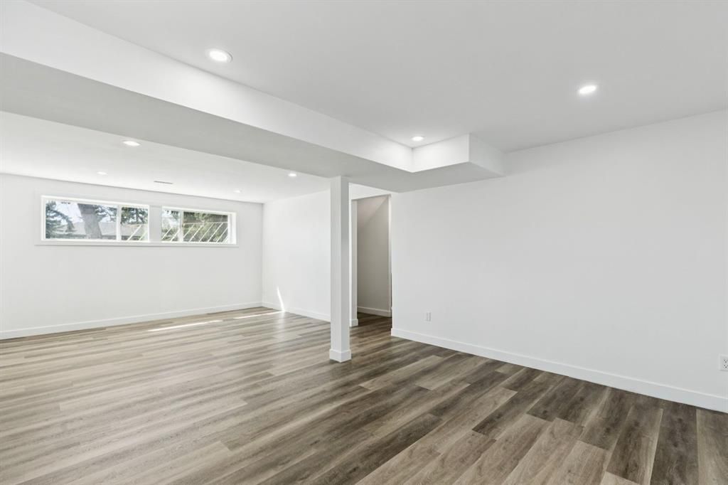 Empty modern room with white walls, wood-look flooring, recessed lights, and a small window.