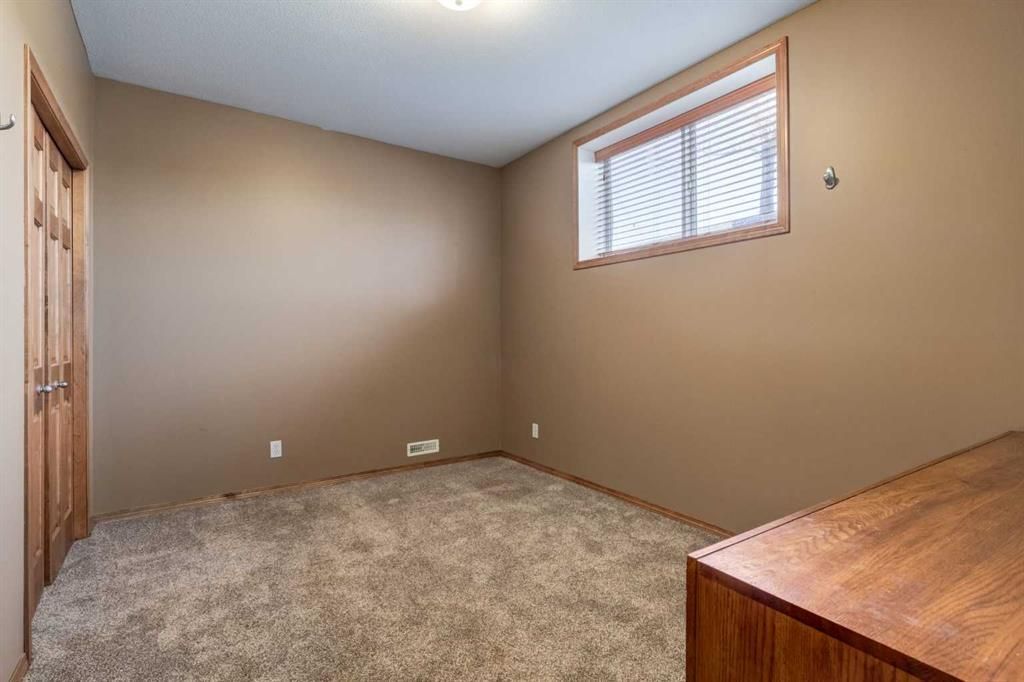 Empty beige bedroom with carpet, a small window, and a wooden dresser corner visible
