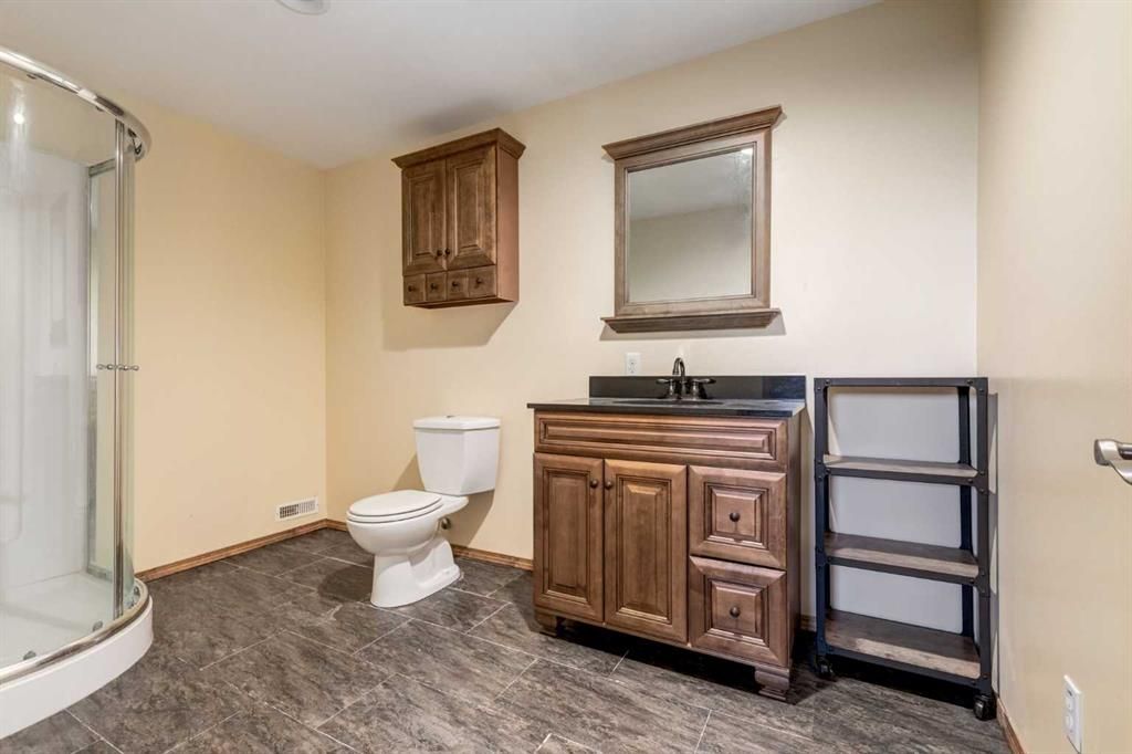 Bathroom with toilet, vanity sink, shower, mirror, and shelving on beige walls.