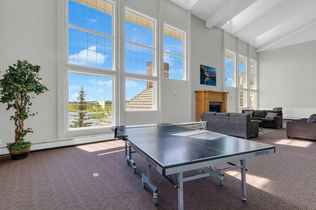 Bright living room with large windows, gray sofas, a fireplace, and a ping-pong table in the center