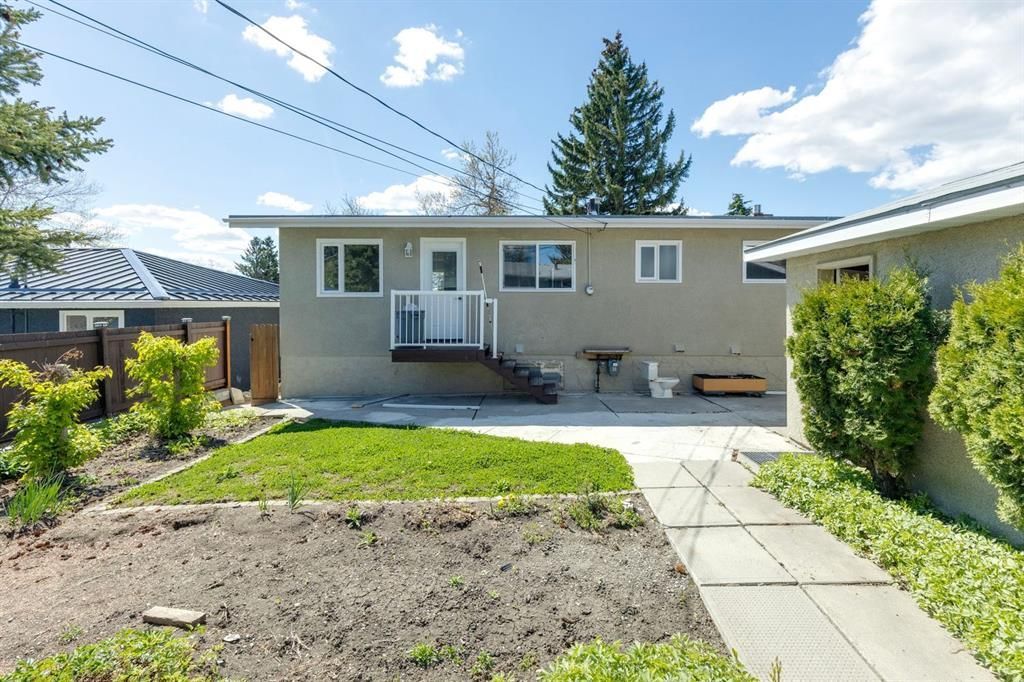 Backyard with a small beige house, front steps, paved path, and trees under a blue sky