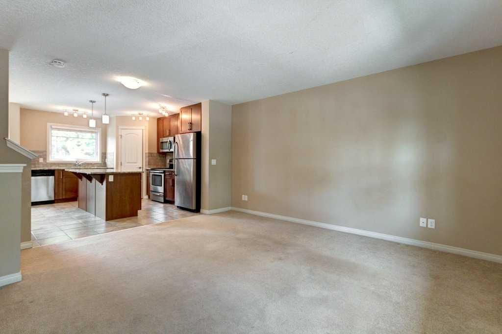 Empty beige living room with carpet, open kitchen, and a refrigerator by the counter