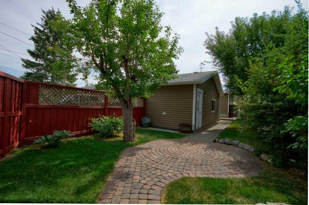Backyard with brick path, green lawn, trees, red fence, and a small shed under cloudy sky