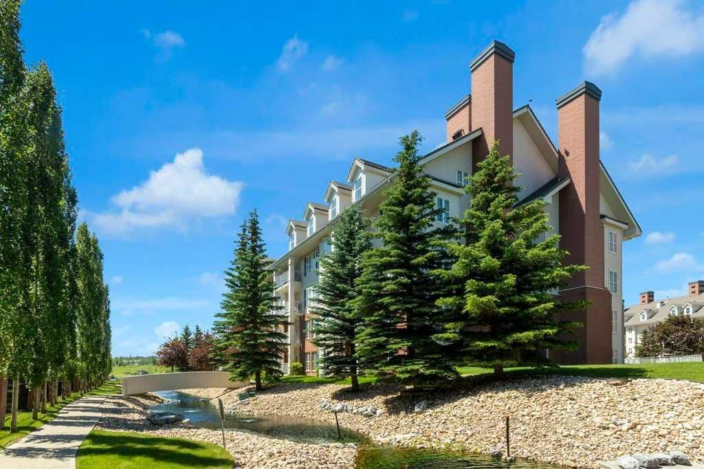 Modern apartment building with tall pine trees, blue sky, and a landscaped walkway beside a rocky path.