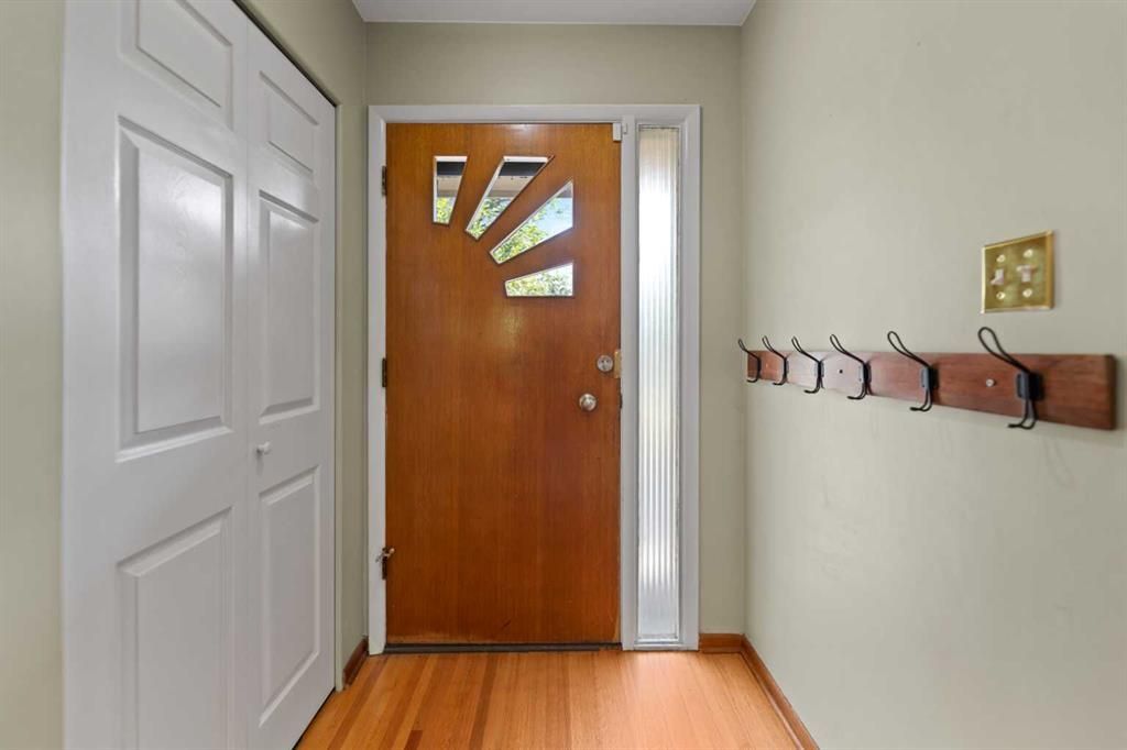 Narrow hallway with white walls, wood floor, coat hooks, and a brown front door with fan-shaped glass panels.