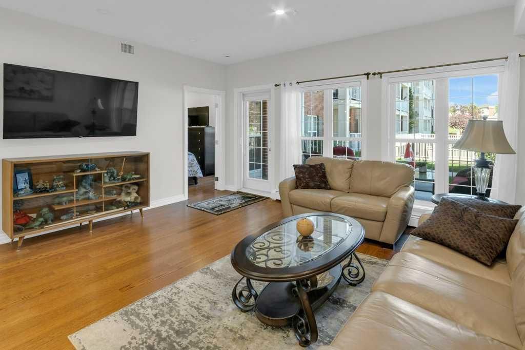 Bright living room with beige sofas, glass coffee table, wall-mounted TV, and fireplace next to large windows.