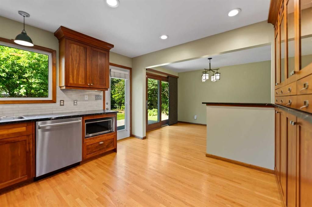 Bright kitchen with wood floors, wooden cabinets, stainless dishwasher, and large windows overlooking greenery