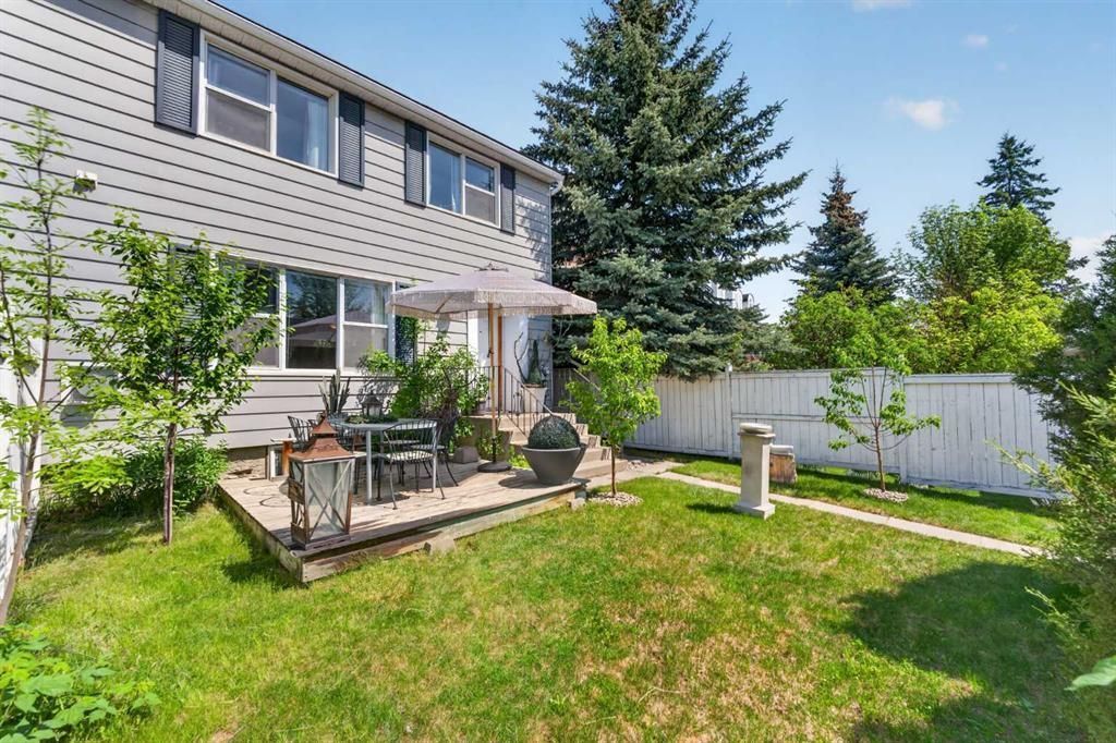 Backyard patio with outdoor seating, green lawn, white fence, and a two-story house in sunlight