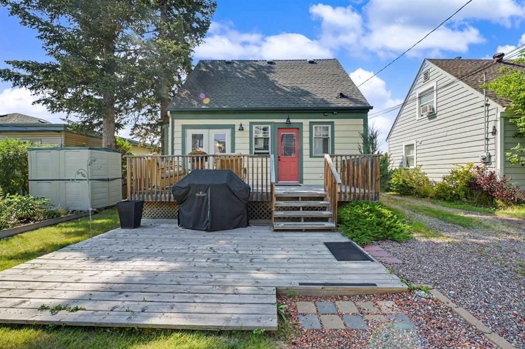 Small house with a front deck, red door, outdoor grill, and paved yard under a blue sky.
