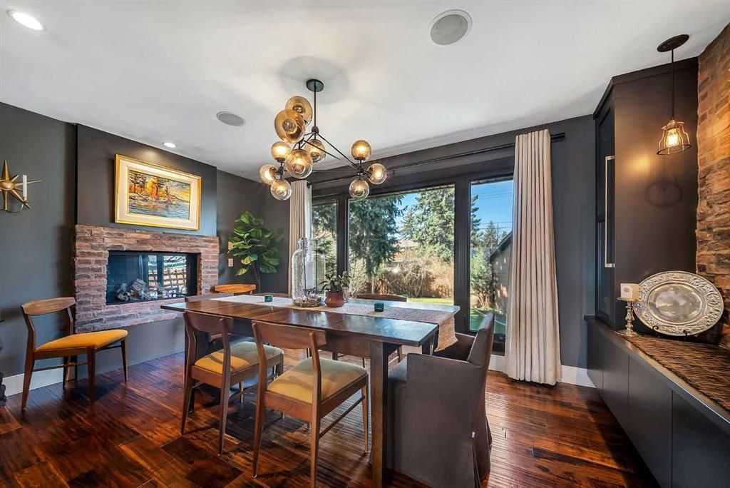 Modern dining room with wooden table, chandelier, fireplace, and large windows overlooking greenery