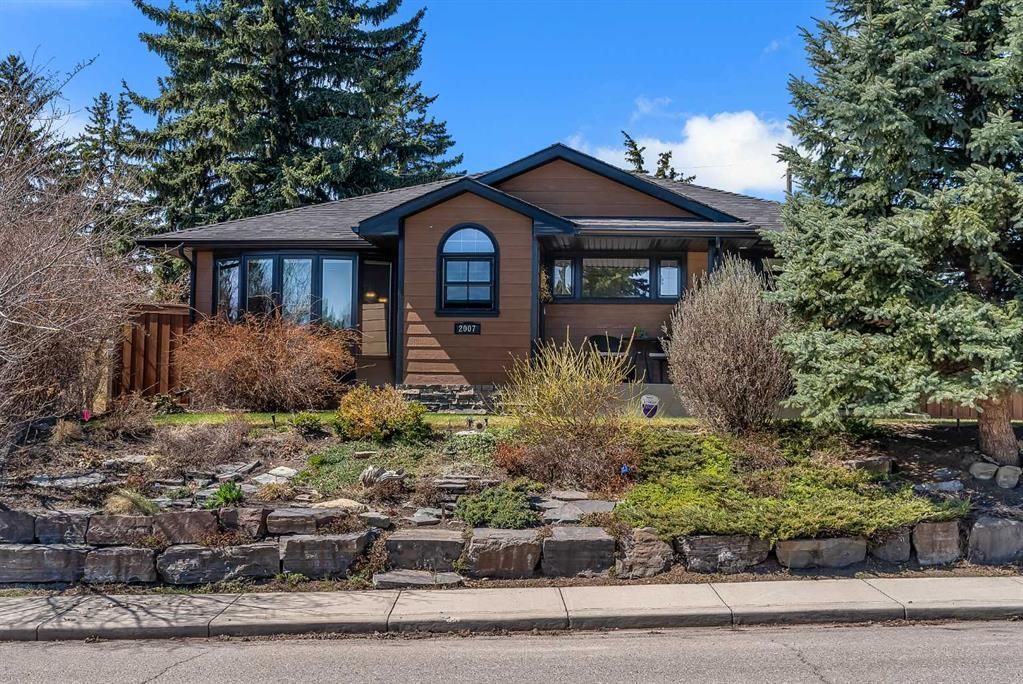Single-story brick house with stone front steps, landscaped yard, and tall evergreen trees under a blue sky