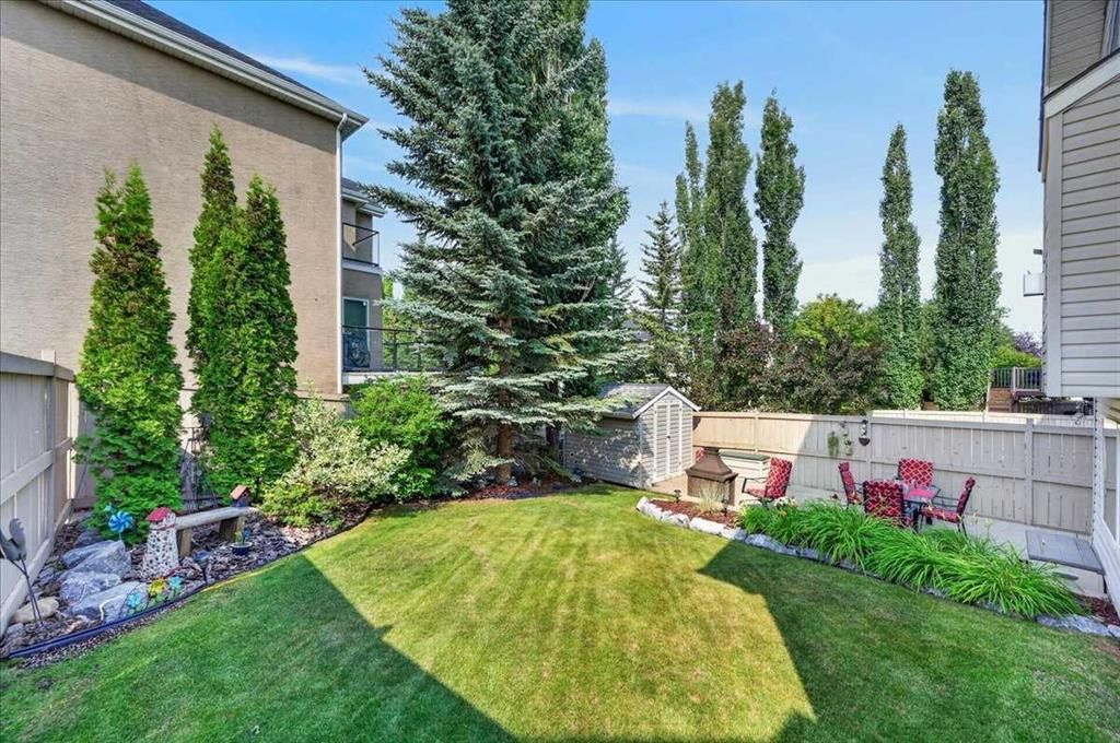 Backyard with green lawn, tall trees, flower beds, and patio seating between two houses