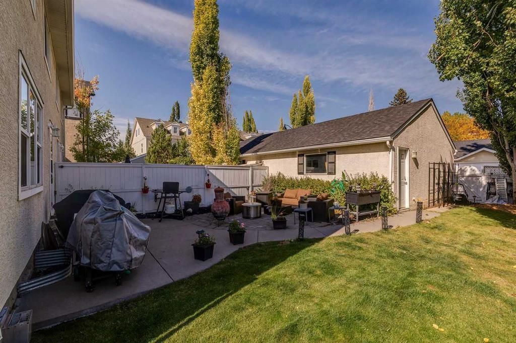 Backyard patio with potted plants, grill, and lawn beside a beige house under a blue sky