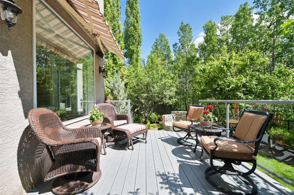 Sunlit patio with wicker chairs, lounge chairs, and lush green trees beside a house.