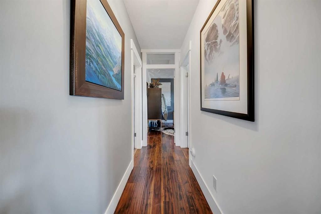 Narrow hallway with hardwood floors and framed artwork on white walls, leading to a room at the end