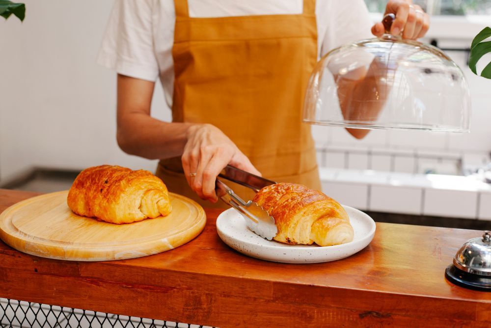 Bakery worker slicing a croissant on a wooden counter beside a covered tray of pastries