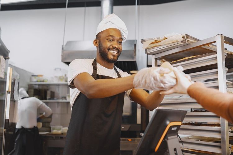 Bakery worker in black apron and white hat handing food to a customer at the counter