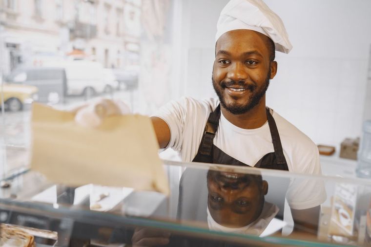 Smiling baker in apron and chef hat handing a pastry across a glass counter